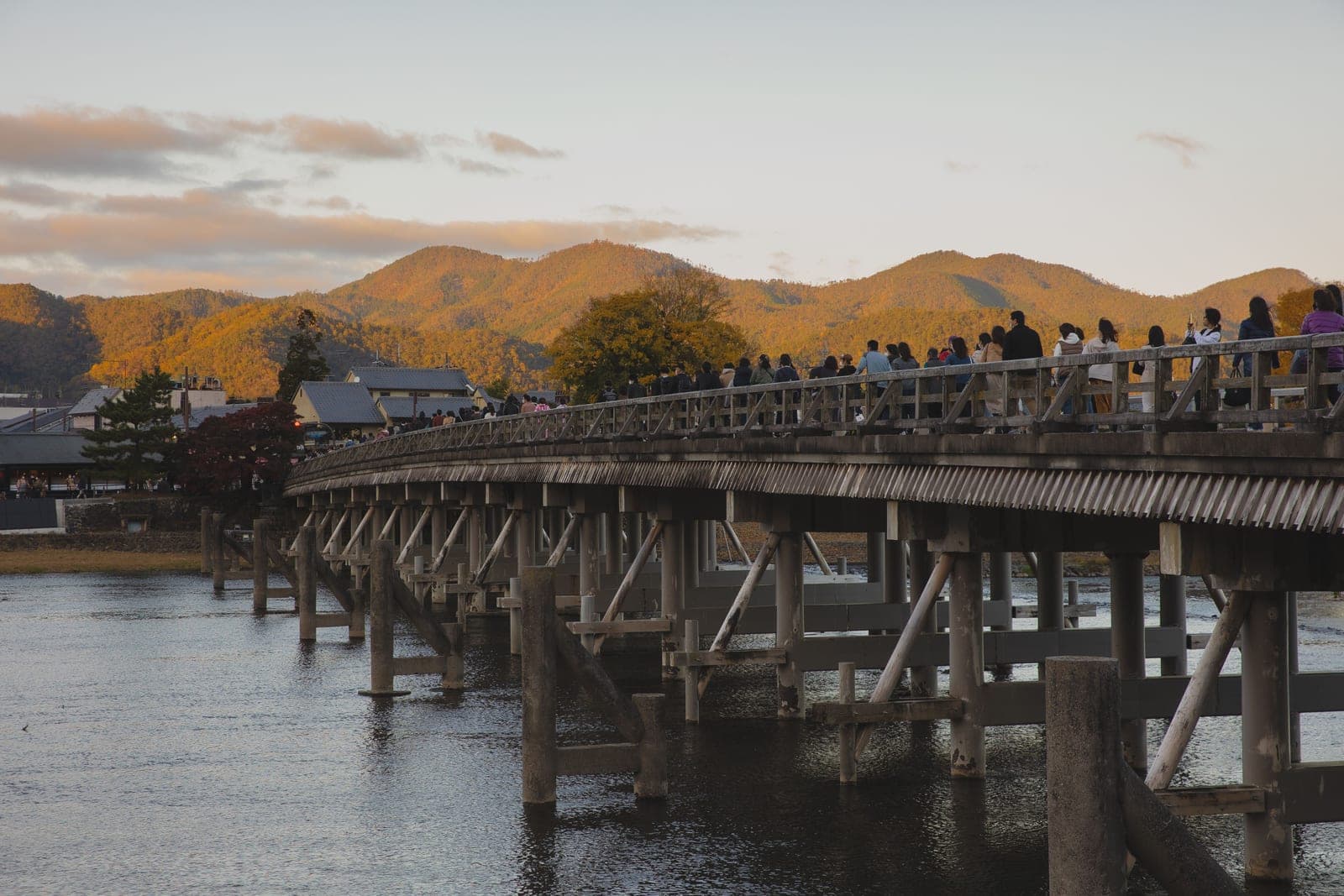 El puente Togetsukyō sobre el río Katsura al atardecer, lleno de visitantes, con las montañas otoñales de Arashiyama al fondo