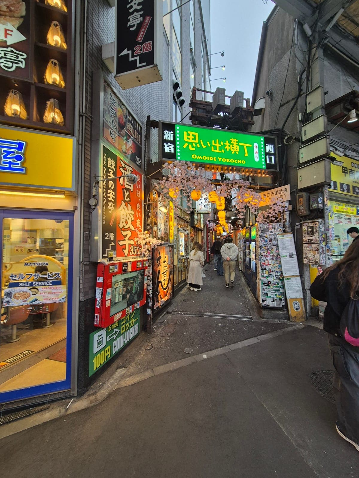 Entrada de Omoide Yokochō en Shinjuku con su letrero verde y cerezos en flor sobre el callejón