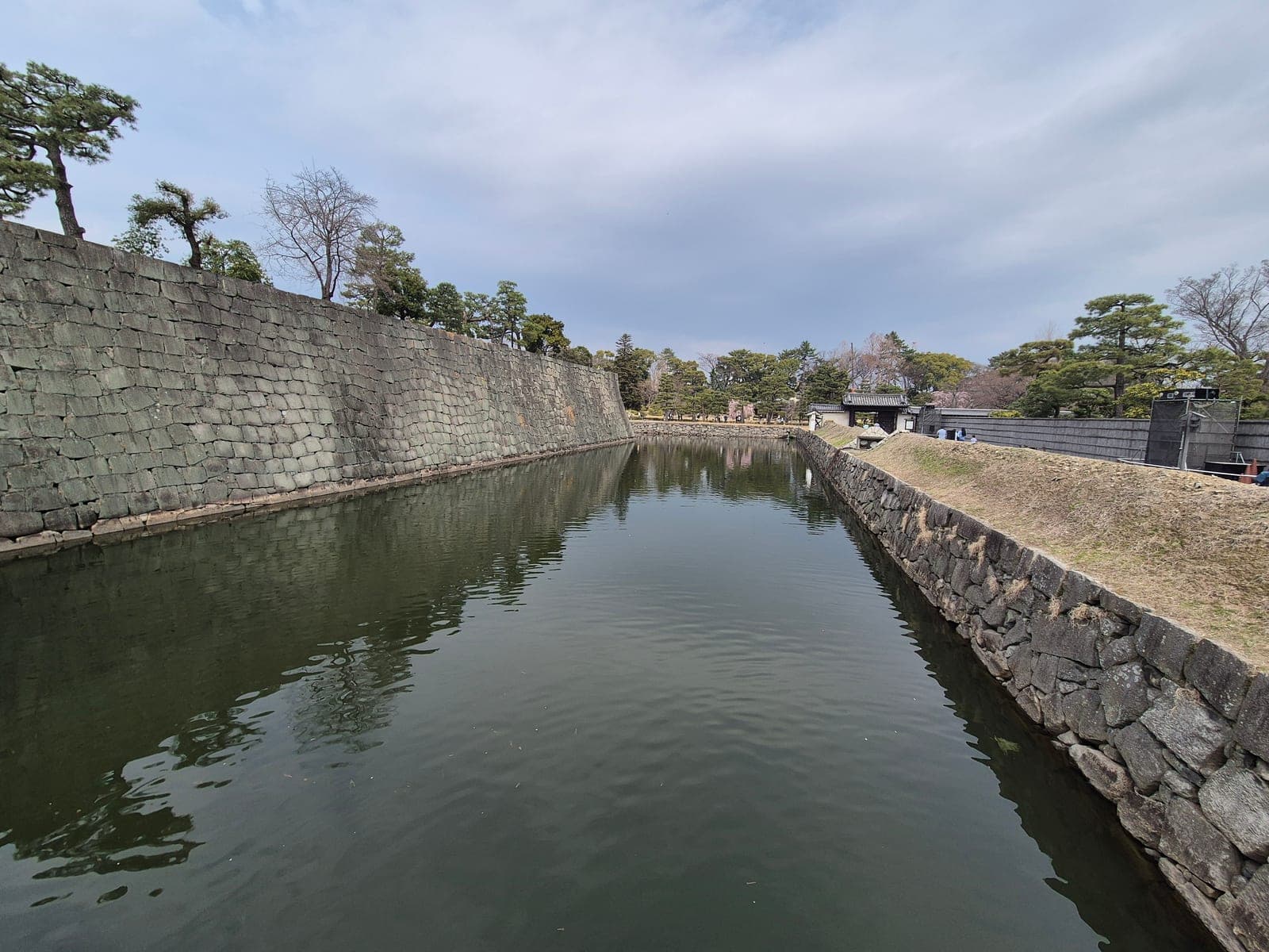 El foso del castillo Nijō entre murallas de piedra, con pinos en la orilla y el agua en calma reflejando el cielo nublado