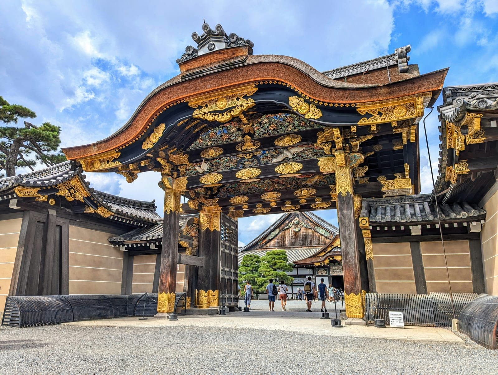 La puerta Karamon del castillo Nijō con sus tallas doradas y policromadas de grullas y flores bajo el techo curvo, y el palacio Ninomaru asomando detrás