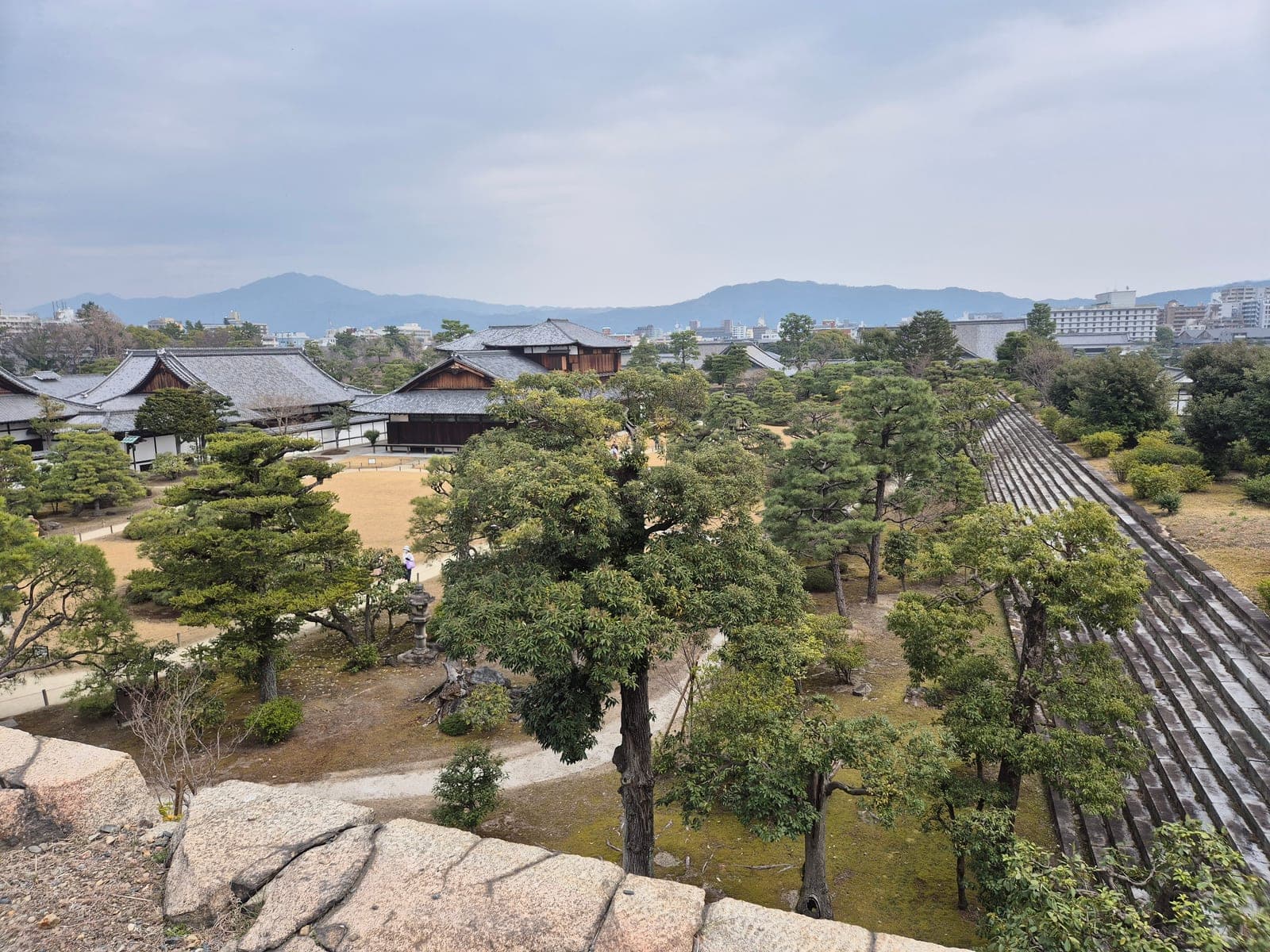 Vista elevada de los jardines del castillo Nijō, con edificios tradicionales, pinos y el perfil de Kioto y las montañas al fondo