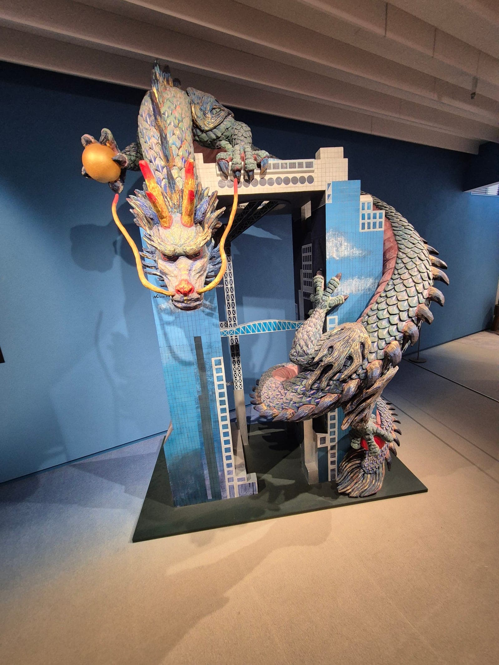 Escultura de colores vivos en el museo Koji Kinutani que muestra un dragón rosa y una figura infantil sobre nubes doradas, junto a un sol estilizado