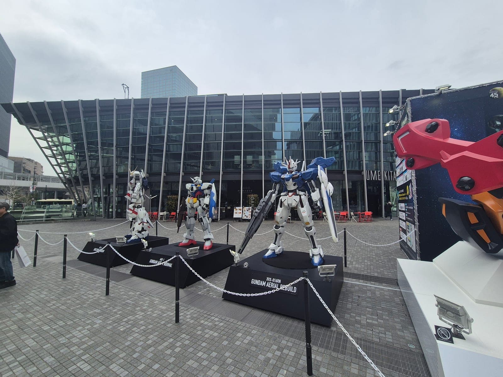 Tres figuras de Gundam a tamaño real montadas sobre plataformas en la plaza de Umekita, Osaka, frente a un edificio de vidrio