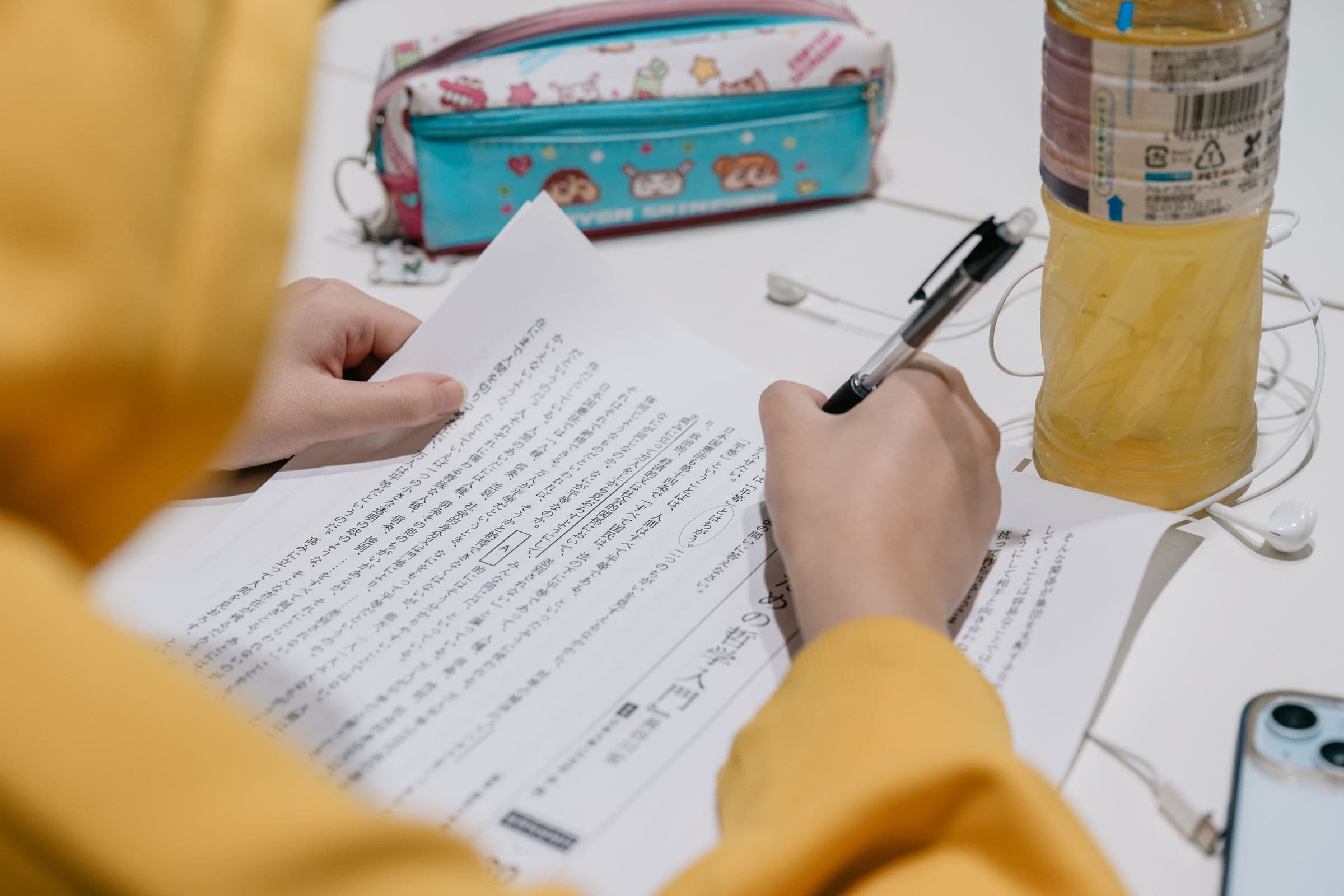 Persona escribiendo con pluma sobre una hoja de estudio con texto en japonés, junto a un estuche y una botella