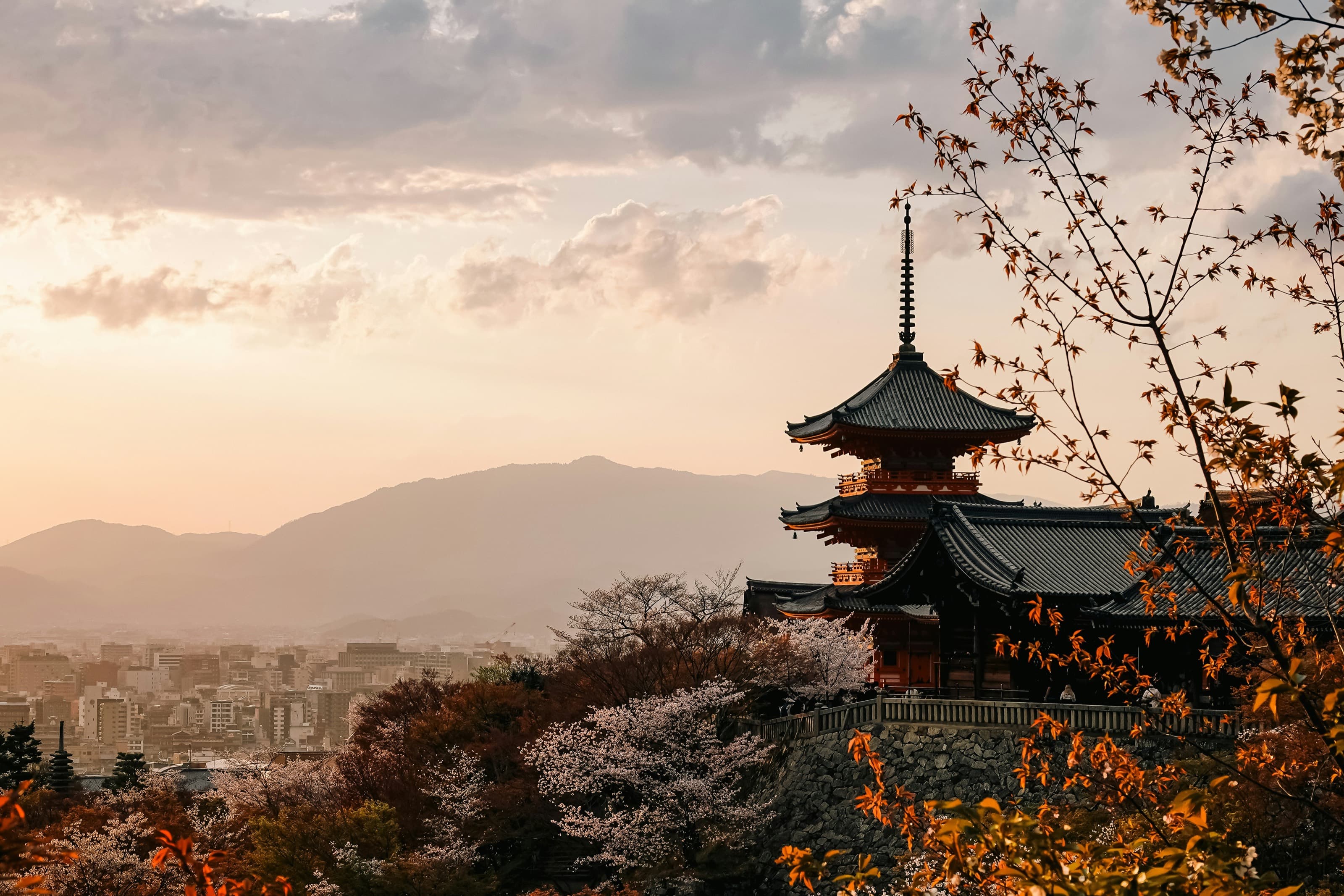 La pagoda de tres pisos del Kiyomizu-dera al atardecer, con cerezos en flor sobre la ladera, la ciudad de Kioto abajo y las montañas al fondo bajo un cielo anaranjado