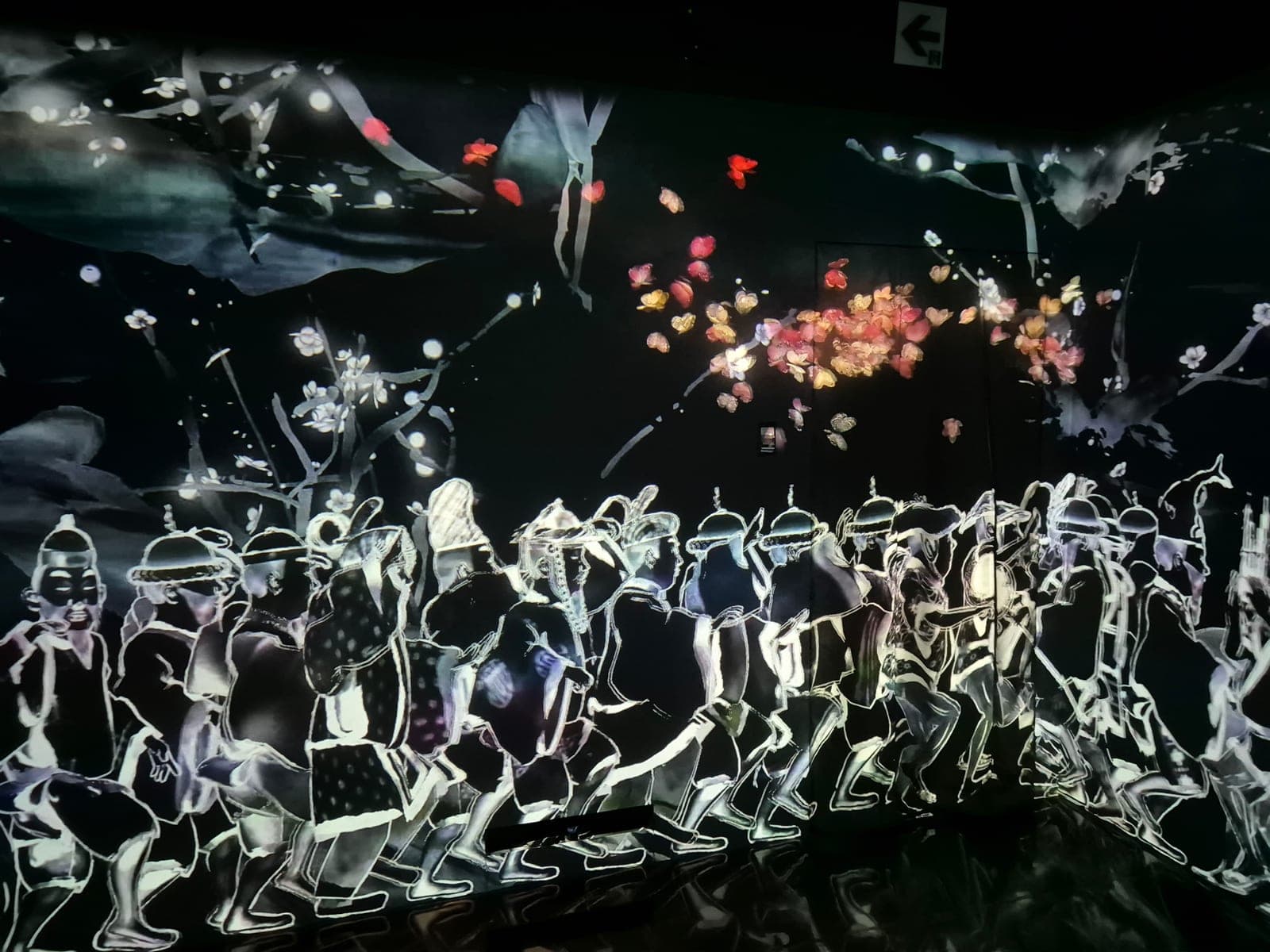 Figuras humanas dibujadas en trazos de luz blanca caminando en fila sobre fondo negro, con pétalos de flores cayendo alrededor, en la instalación Walk, Walk, Walk de teamLab Borderless