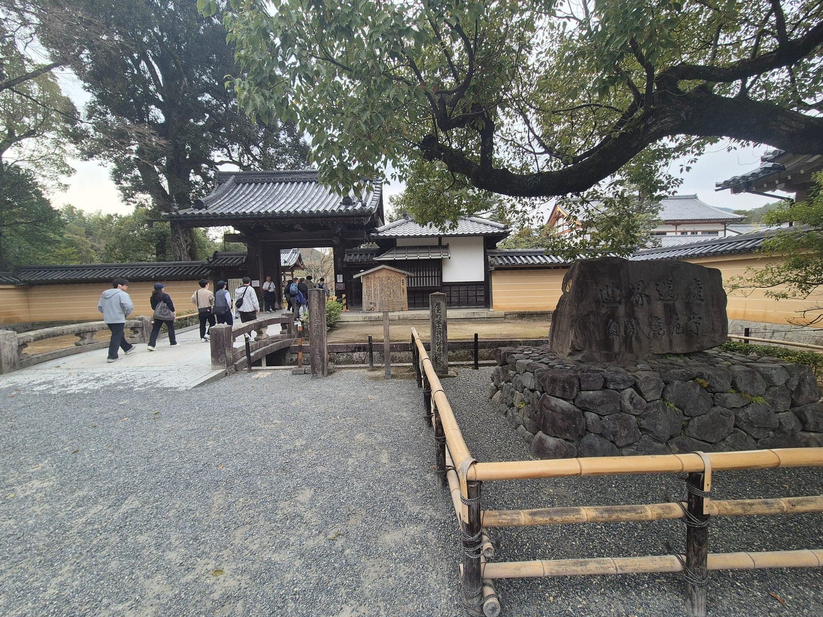 Entrada del templo Kinkaku-ji en Kioto, con la puerta tradicional, un monumento de piedra con inscripciones y visitantes cruzando el puente de acceso