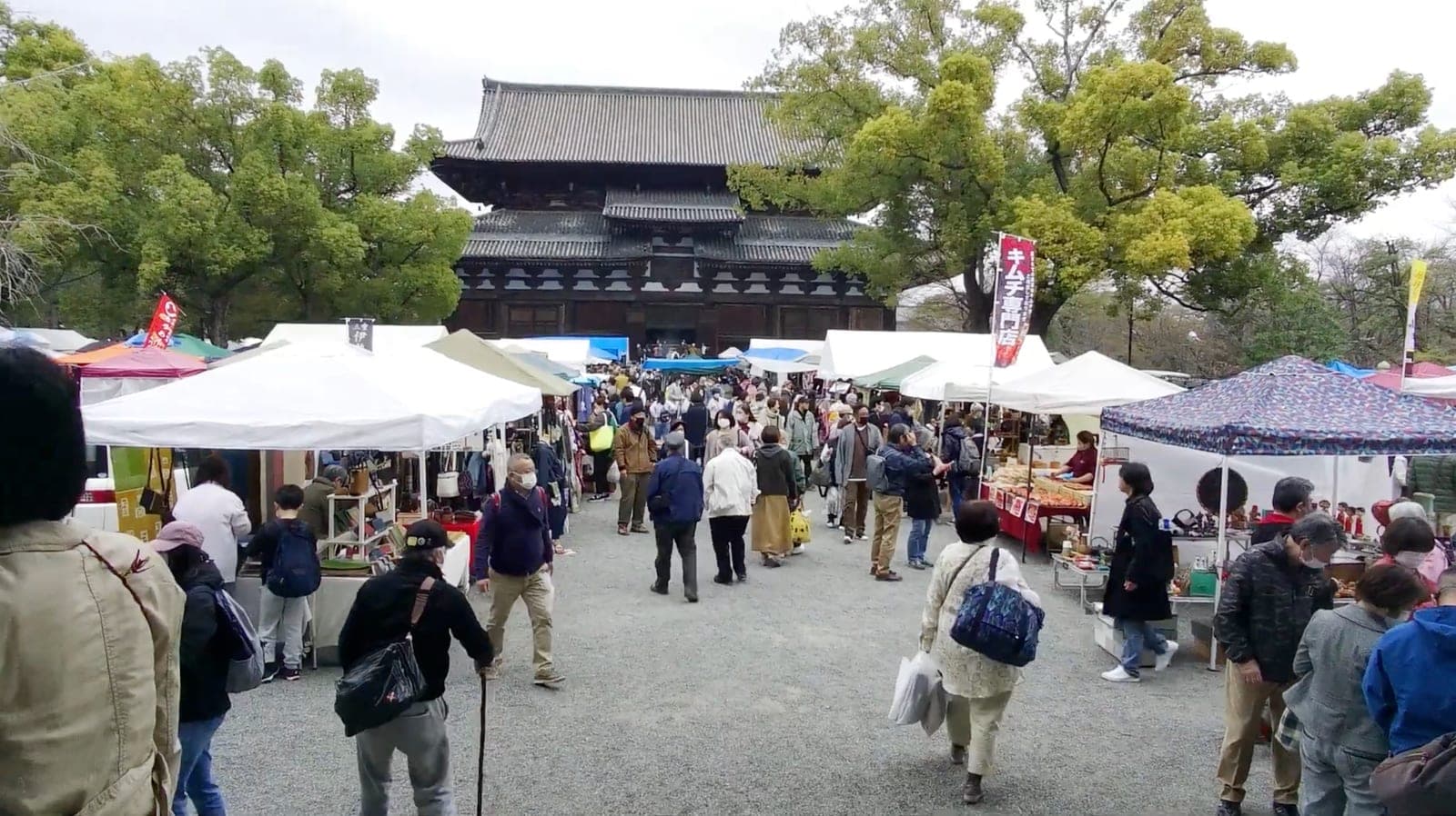 Puestos con toldos del mercado Kōbō-ichi llenos de gente en los patios del templo Tō-ji, con el Kōdō de fondo