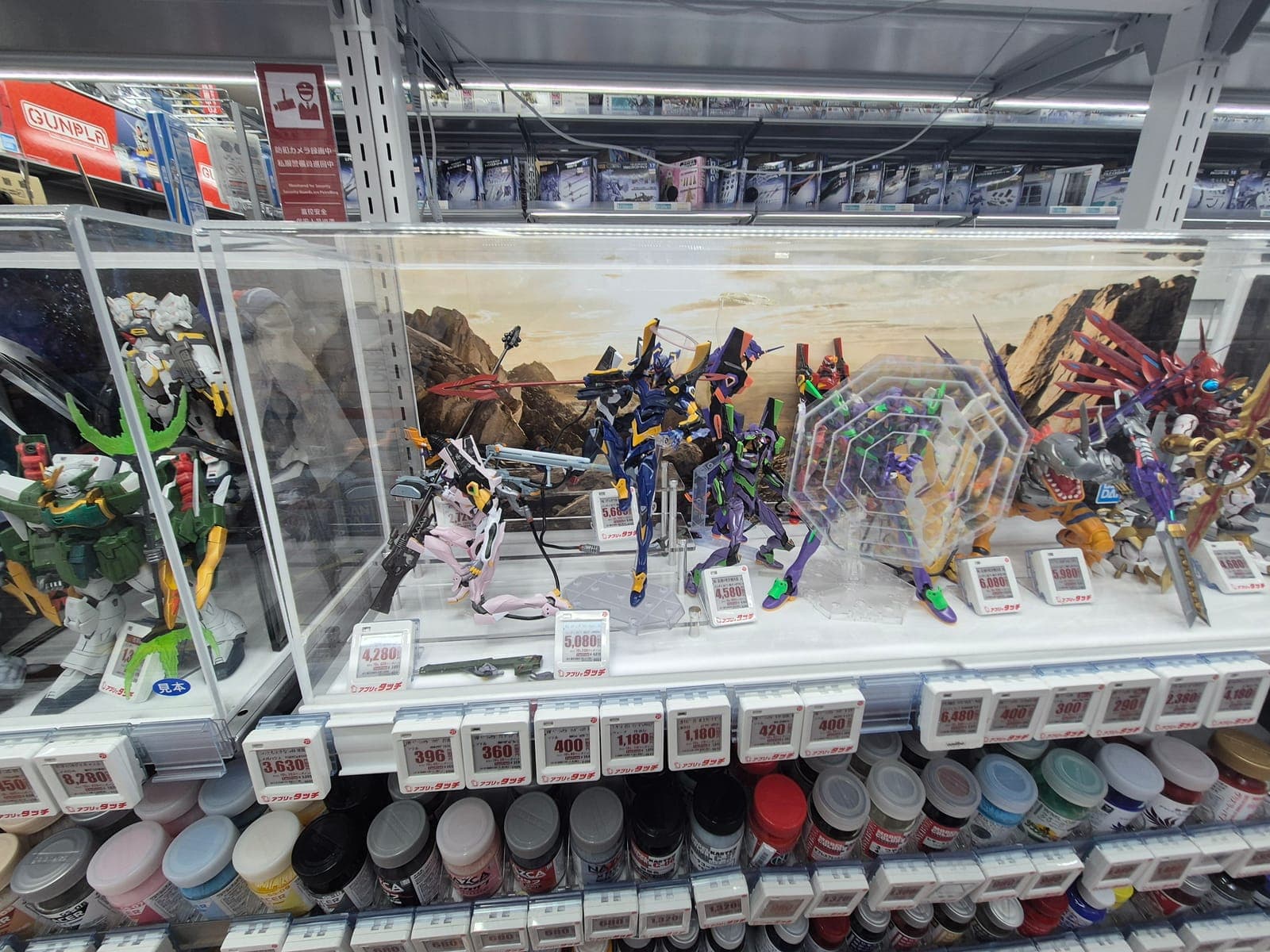 Vitrina con kits de modelismo de Evangelion posados en escena, con precios y frascos de pintura en los estantes