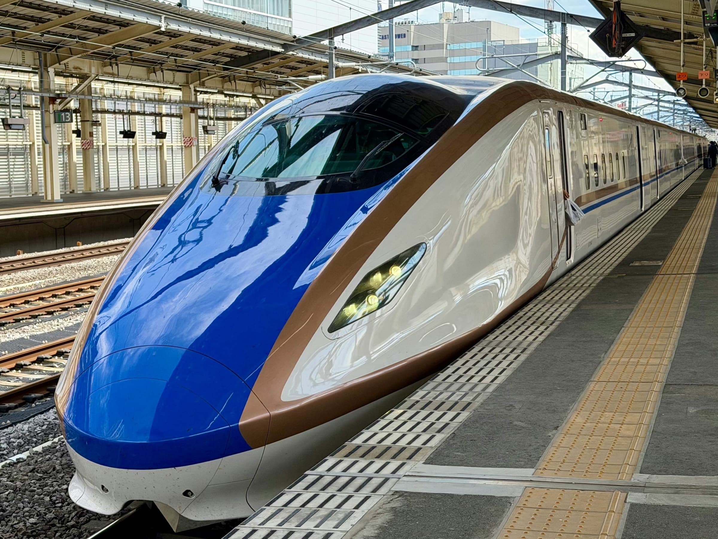 Tren bala shinkansen de la serie E7/W7 detenido en el andén de una estación, con edificios de la ciudad al fondo