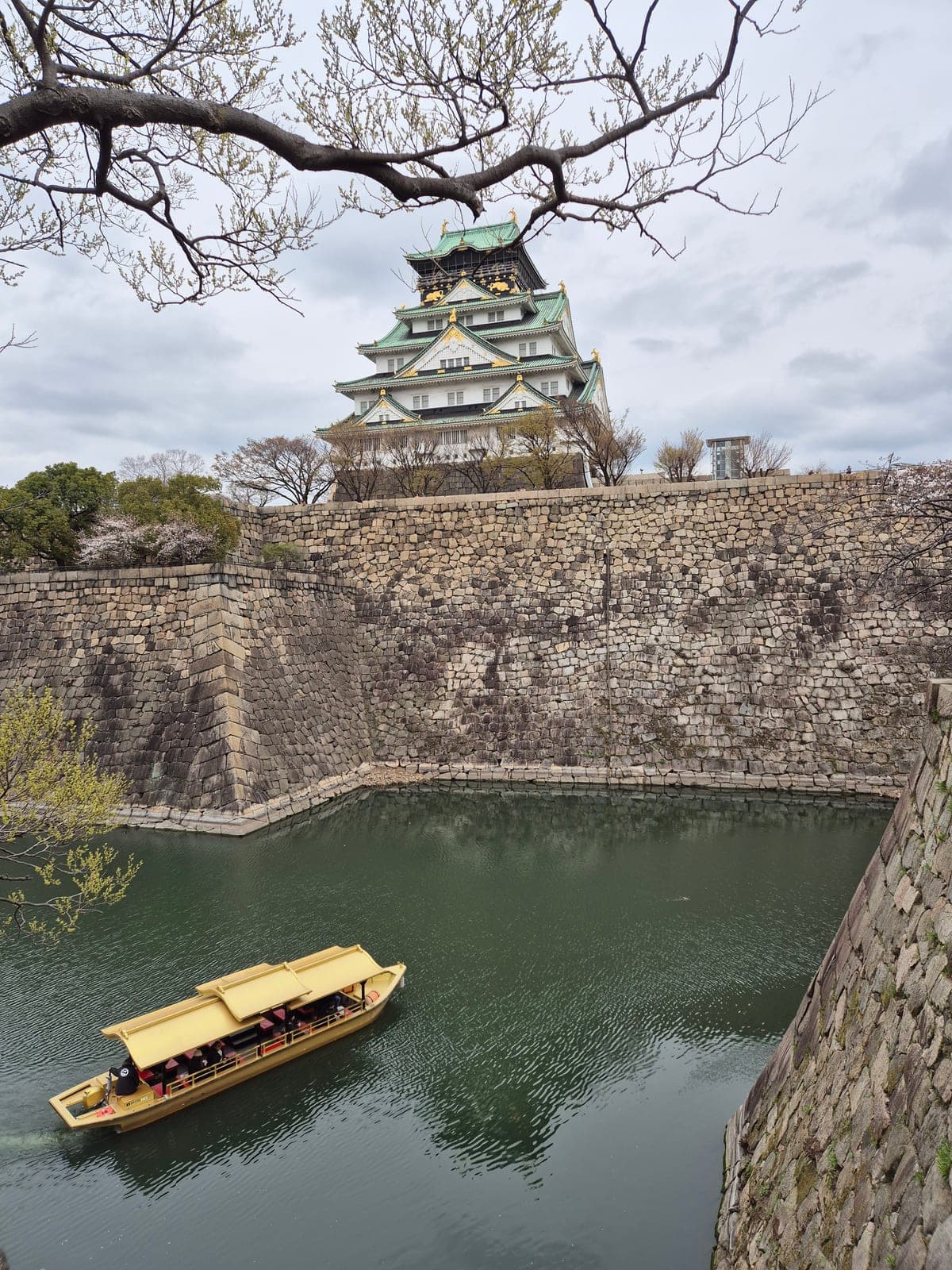 Un barco gozabune dorado navegando por el foso del Castillo de Osaka, con la torre principal y las murallas de piedra al fondo