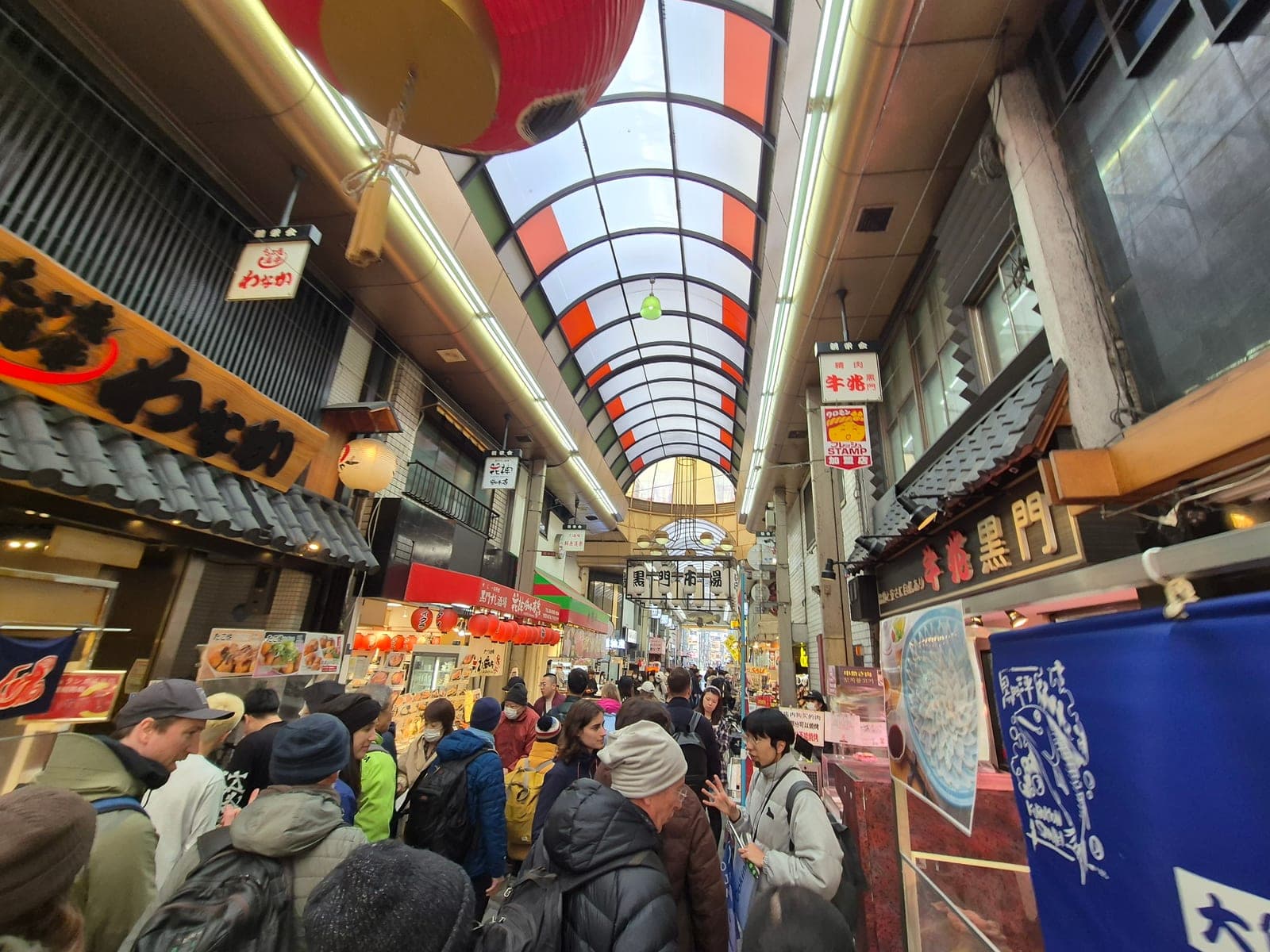 Pasillo techado de Kuromon Ichiba en Osaka con un farol rojo colgante, letreros de tiendas y visitantes caminando entre los puestos