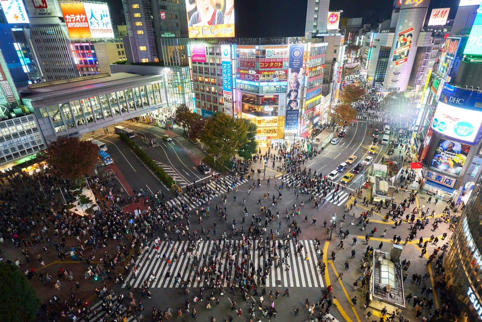 El cruce de Shibuya visto desde arriba de noche, con cientos de peatones cruzando en todas direcciones entre pantallas y letreros iluminados