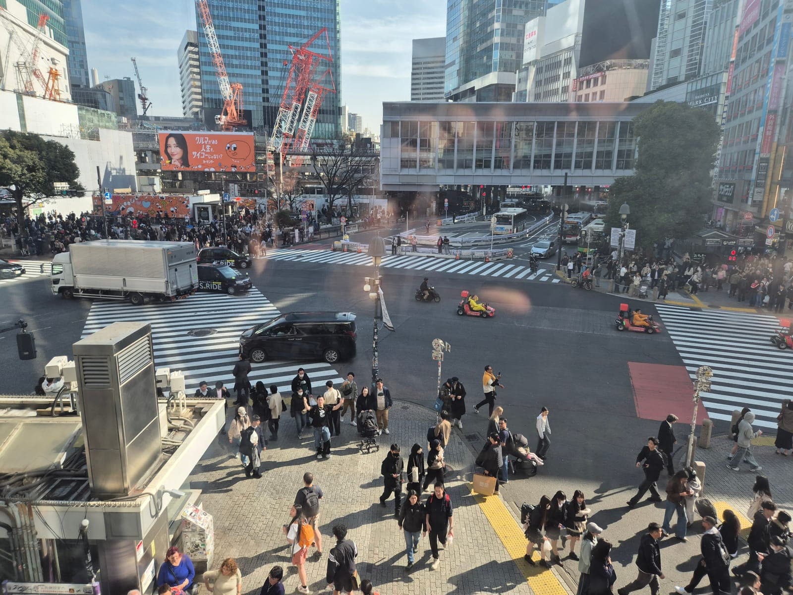 Vista elevada del cruce peatonal de Shibuya con la multitud cruzando en todas direcciones entre grúas de construcción y rascacielos