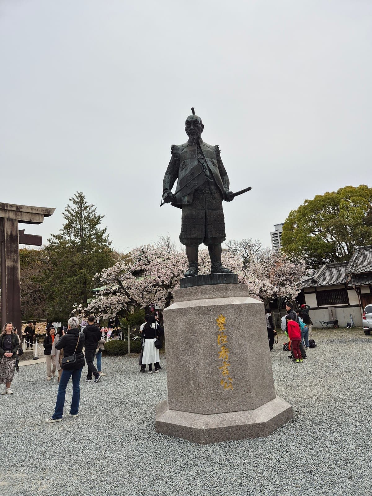Estatua de bronce de Toyotomi Hideyoshi con armadura samurái completa sobre un pedestal de piedra, rodeada de flores de cerezo en Hokoku Shrine