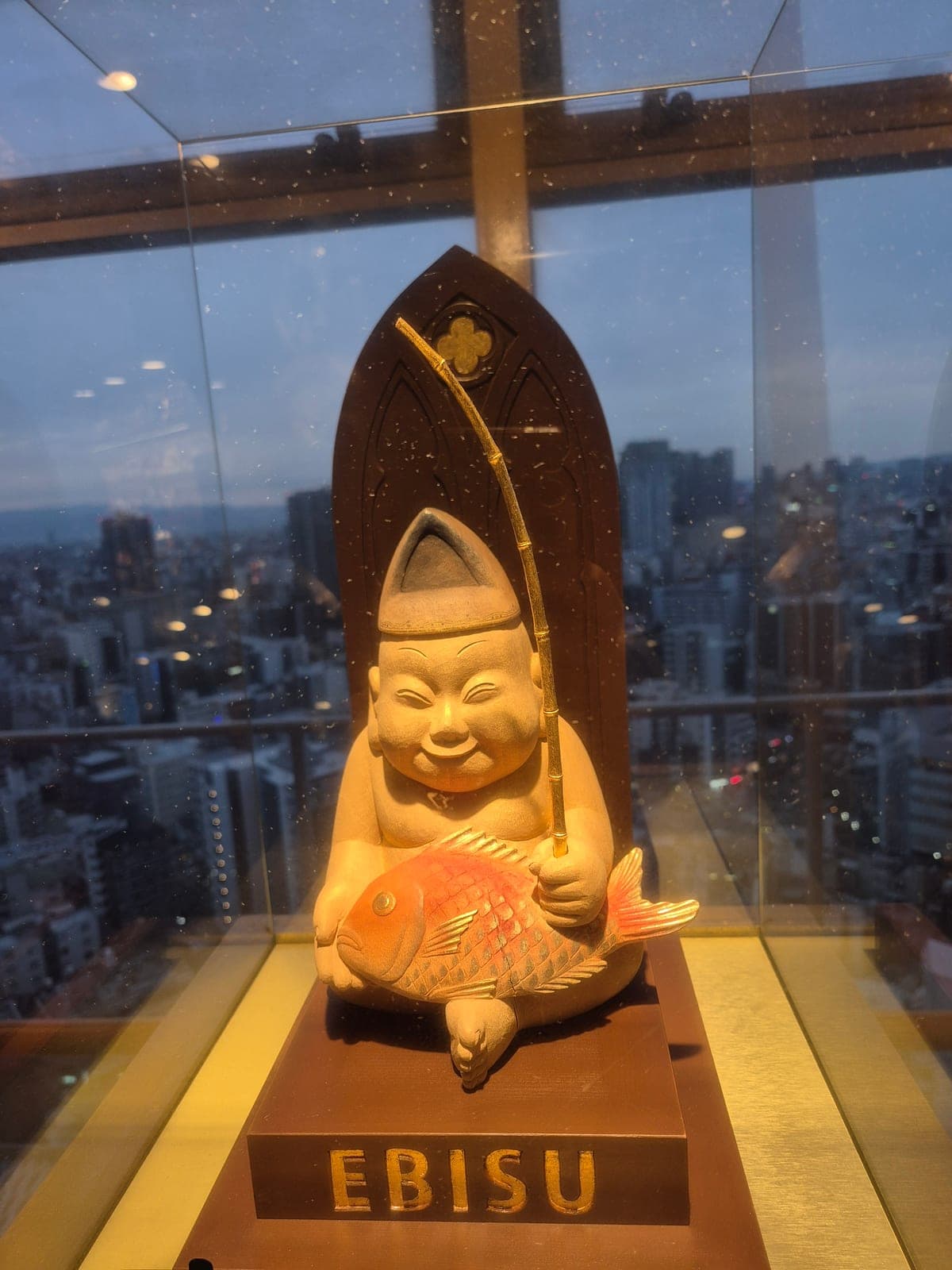 Estatua dorada de Ebisu sosteniendo una vara de pescar y un pez, dentro de la torre Tsutenkaku, con la ciudad de Osaka de fondo a través del cristal