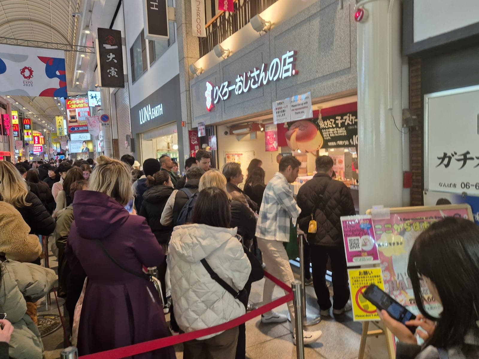 Fila de turistas y locales frente a la tienda Rikuro Ojisan en Shinsaibashi-suji, Osaka, con un letrero grande de un cheesecake recién horneado