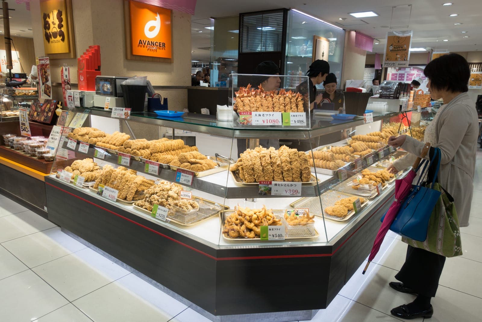 Vitrina de una depachika con bandejas de tonkatsu, croquetas y camarones empanizados, y una clienta viendo los precios
