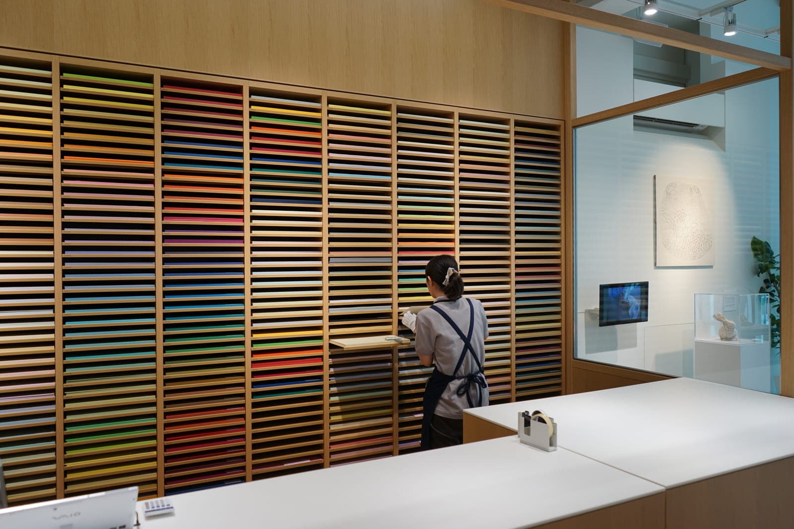 Pared de papeles de colores organizados por tono dentro de Itoya, con una empleada acomodando la mercancía