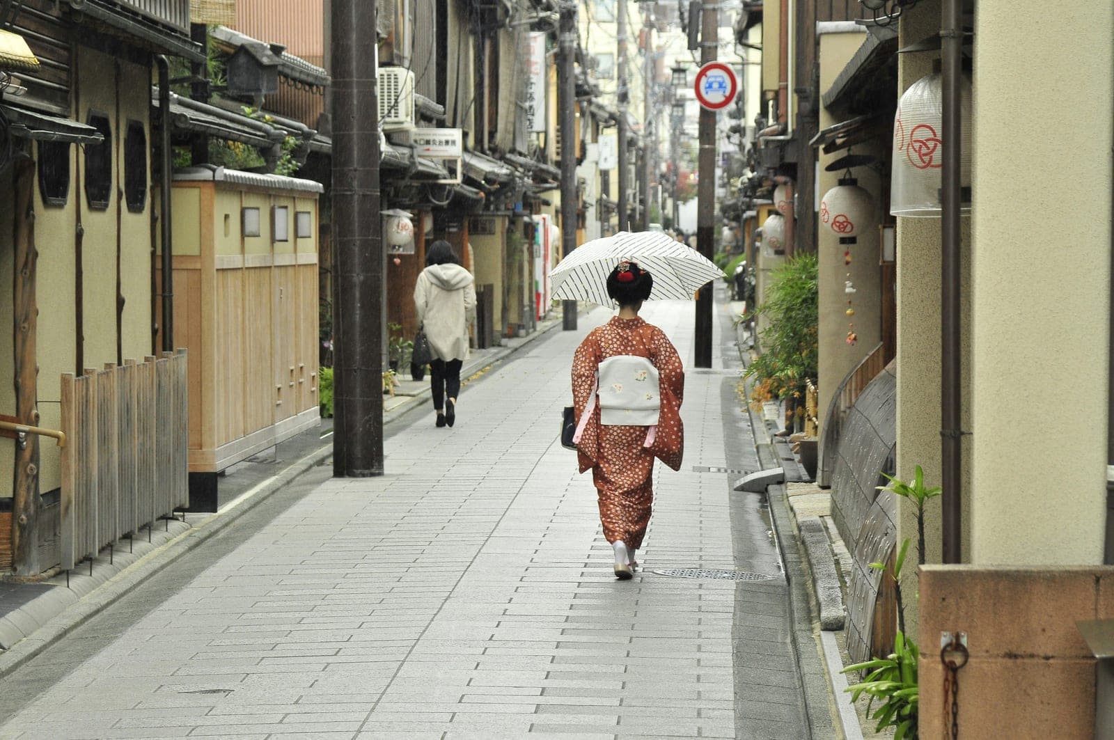 Una maiko caminando de espaldas bajo una sombrilla por una calle tradicional de Gion, con kimono y obi claros