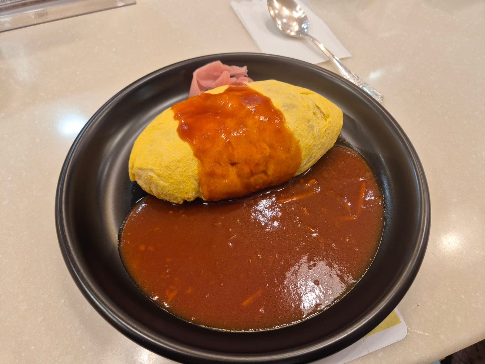 Un omurice servido en un plato negro en Hokkyokusei, Osaka, con la tortilla de huevo bañada en salsa demi-glace y un poco de jengibre encurtido al lado