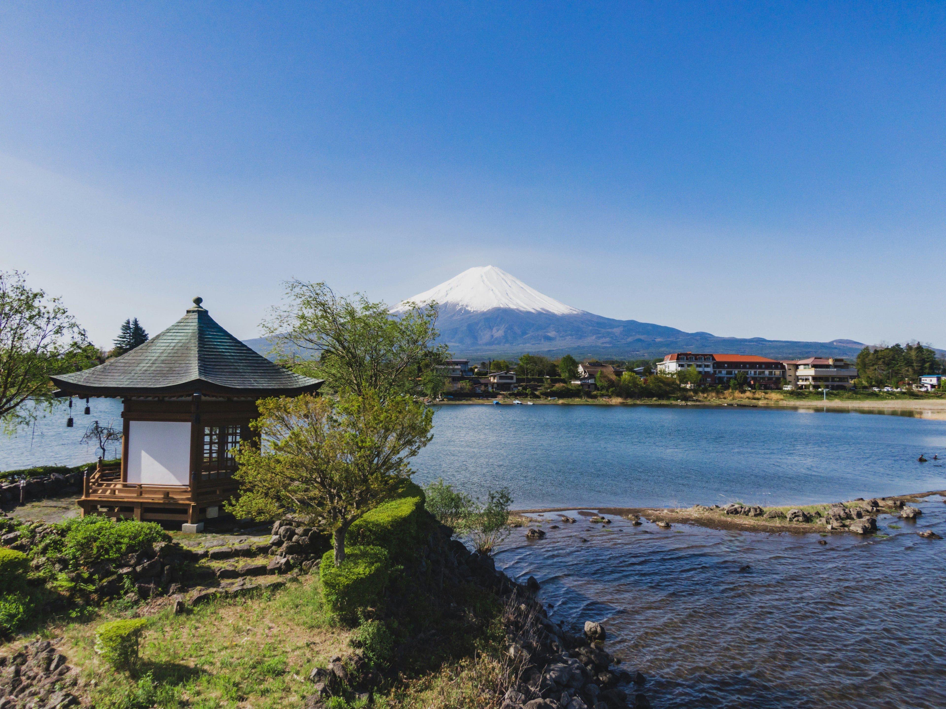 Un pabellón de madera con techo verde en la orilla del lago Kawaguchiko, con el Monte Fuji nevado al fondo y el cielo despejado