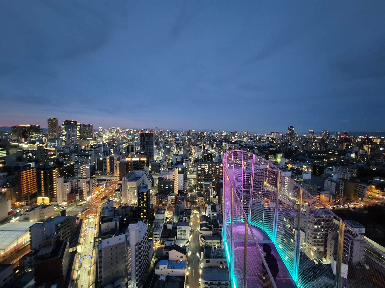 La pasarela iluminada del Tip de la torre Tsutenkaku de noche, con un visitante caminando hacia la punta y las luces de Osaka extendiéndose abajo