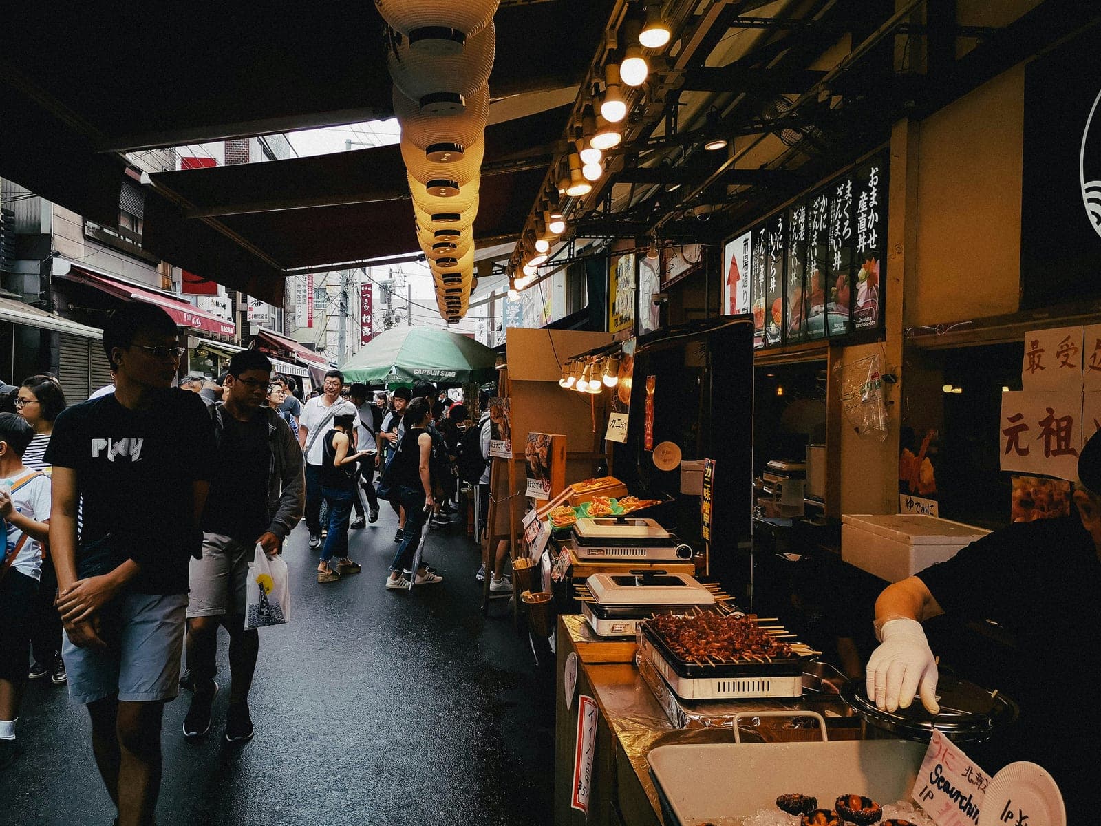 Puesto callejero de Tsukiji con brochetas de mariscos asándose sobre planchas eléctricas, junto a un letrero rojo y gente pasando por el callejón