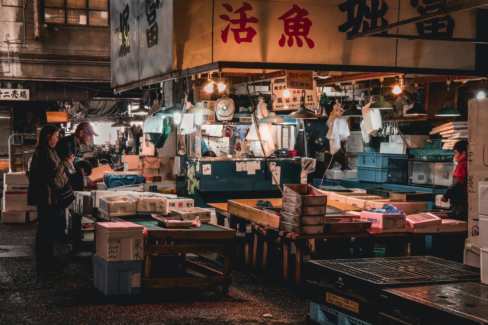 Interior de un puesto de pescado en Tsukiji al anochecer, con letreros japoneses colgantes, lámparas encendidas y clientes revisando la mercancía