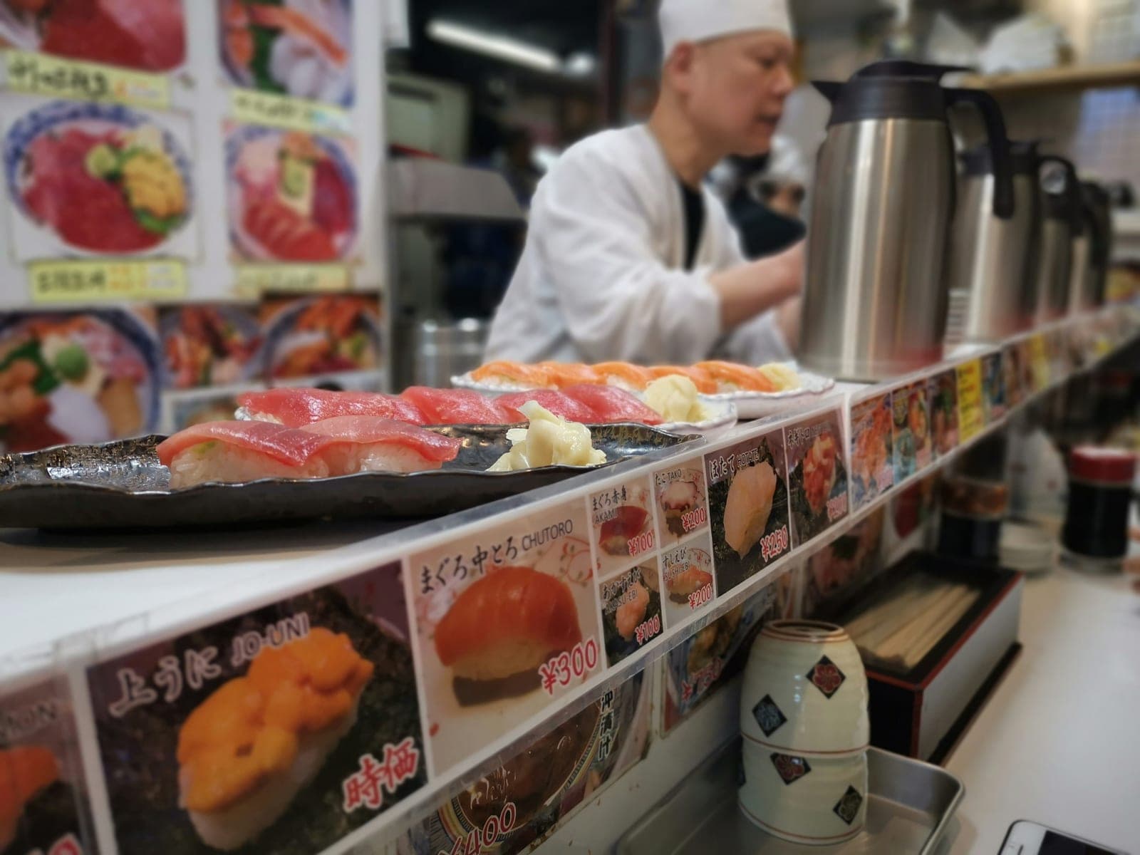Barra de un puesto de sushi en Tsukiji con piezas de atún y salmón recién preparadas sobre platos, y un menú con precios por pieza