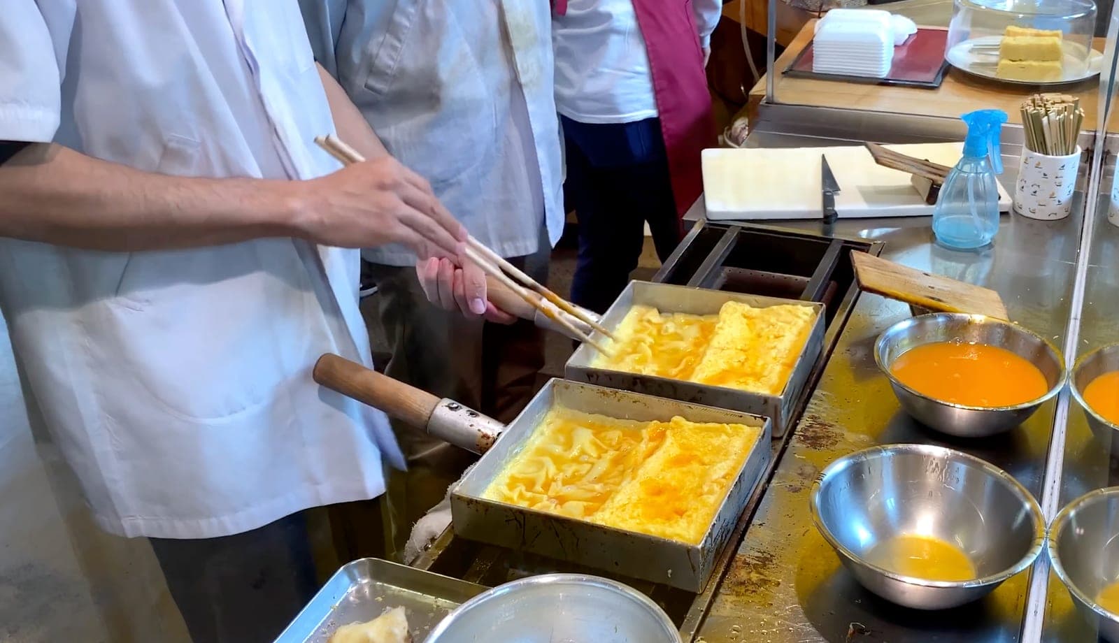 Cocinero preparando tamagoyaki en Tsukiji, doblando capas de huevo con palillos largos sobre moldes rectangulares de metal, con tazones de mezcla de huevo alrededor