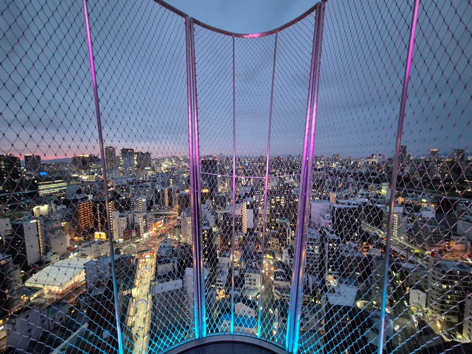 La terraza al aire libre Tenbo Paradise de la torre Tsutenkaku al atardecer, con la malla metálica iluminada de rosa y azul y las luces de Osaka al fondo