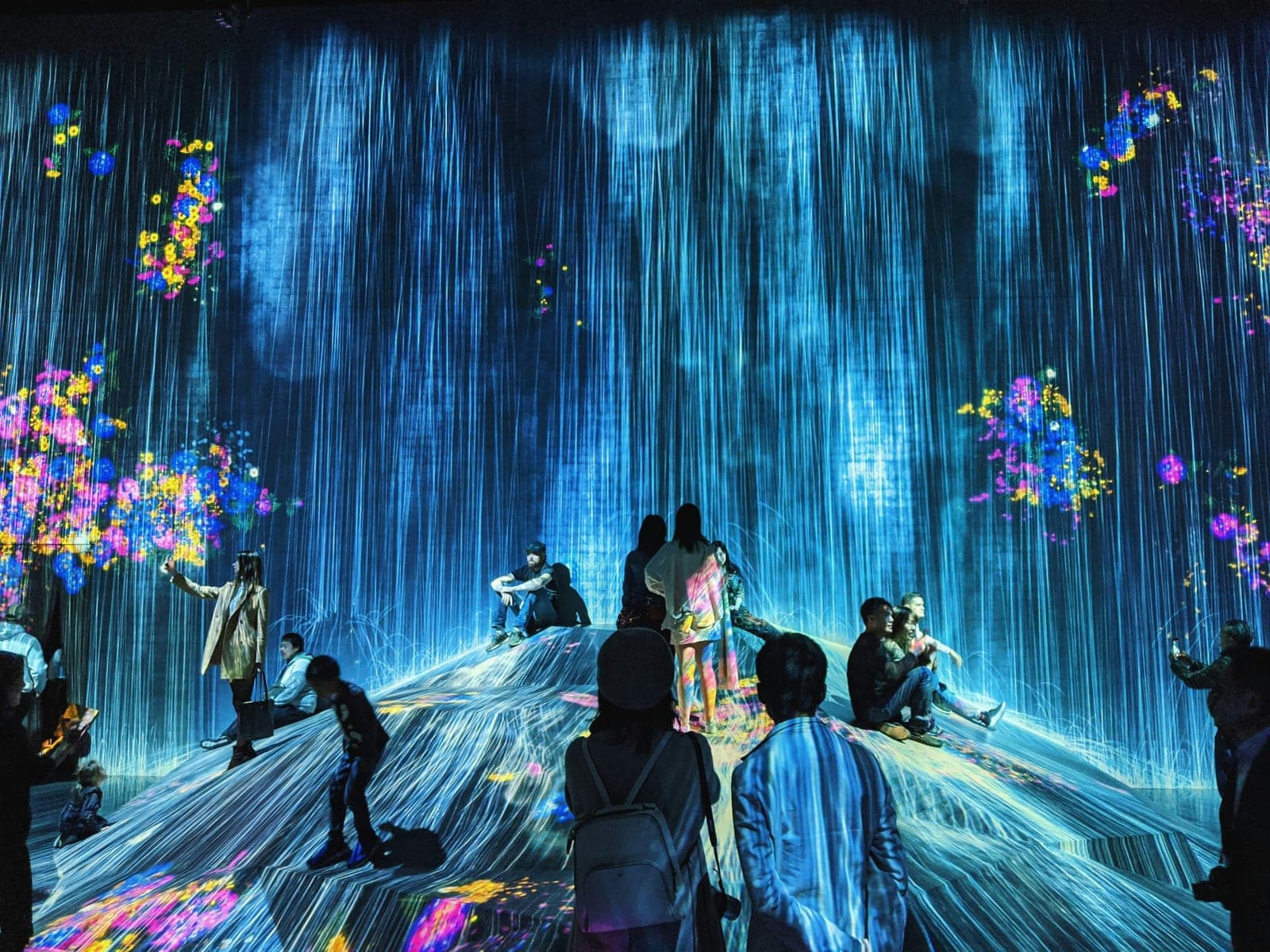 Visitantes sobre un montículo dentro de una sala de teamLab Borderless, rodeados de líneas de luz azul como una cascada
