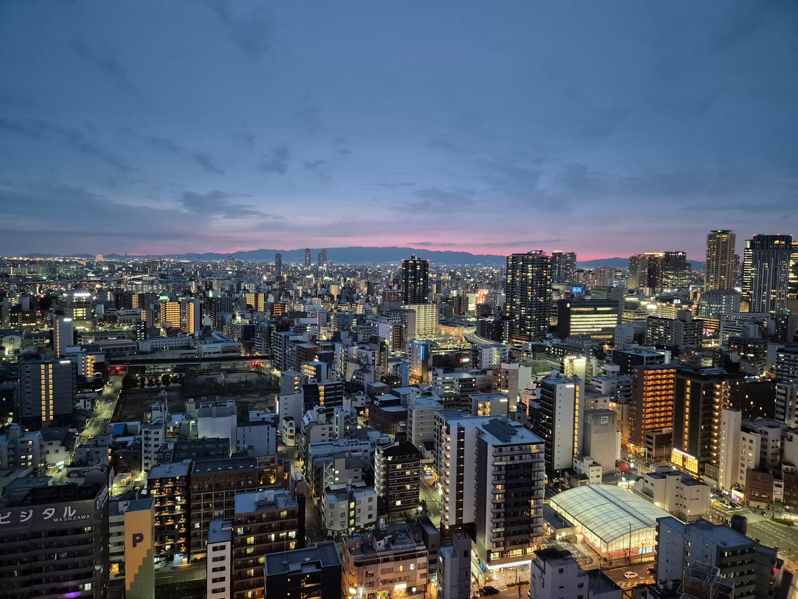 Las luces de Osaka al anochecer desde la torre Tsutenkaku, con los últimos tonos rosas del atardecer sobre las montañas al fondo