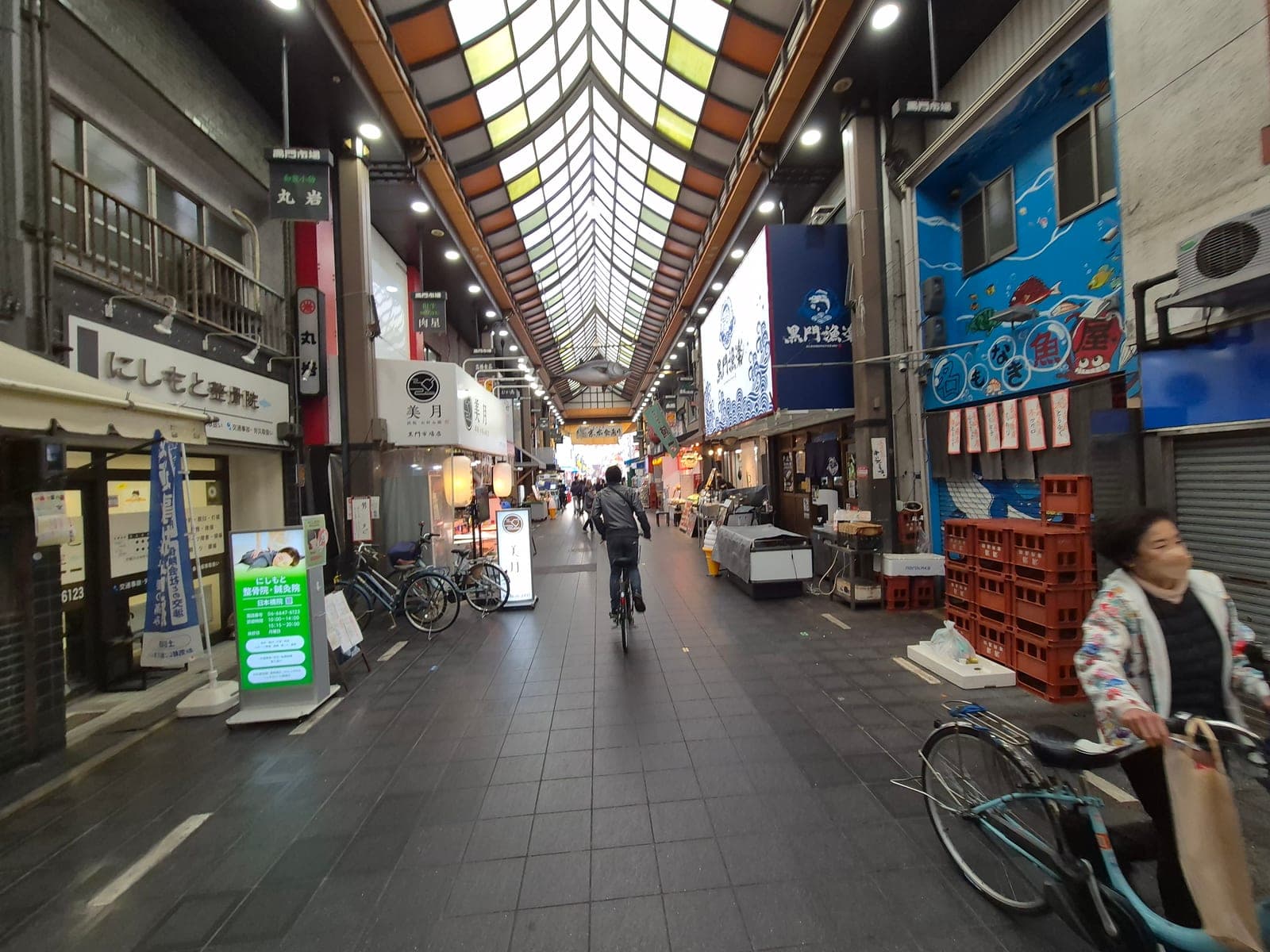 Pasillo de Kuromon Ichiba con pescaderías y tiendas a los lados, bicicletas estacionadas y un techo de tragaluz iluminando el corredor