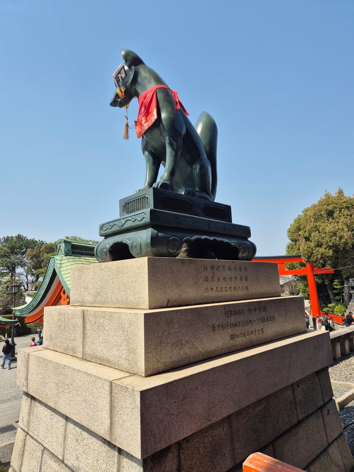 Estatua de zorro kitsune de piedra oscura con babero rojo, sosteniendo una llave en la boca, junto a un torii en Fushimi Inari Taisha