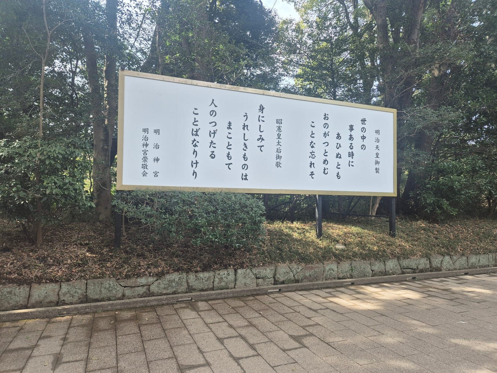 Placa blanca con caligrafía japonesa vertical que muestra dos poemas waka, uno del emperador Meiji y otro de la emperatriz Shōken, junto a un camino del santuario entre árboles