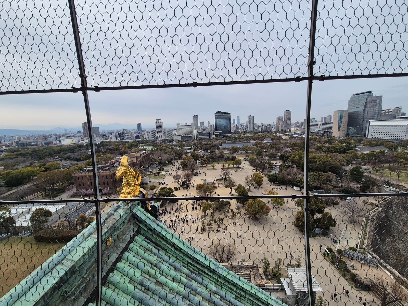 Un shachihoko dorado en la esquina del tejado del Castillo de Osaka, visto de cerca desde el mirador del octavo piso a través de la malla de seguridad, con el horizonte de la ciudad al fondo