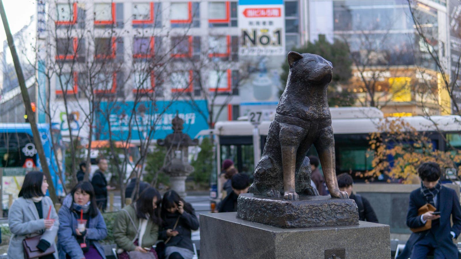 La estatua de bronce de Hachikō en Shibuya, de cerca, con gente y camiones alrededor de la plaza