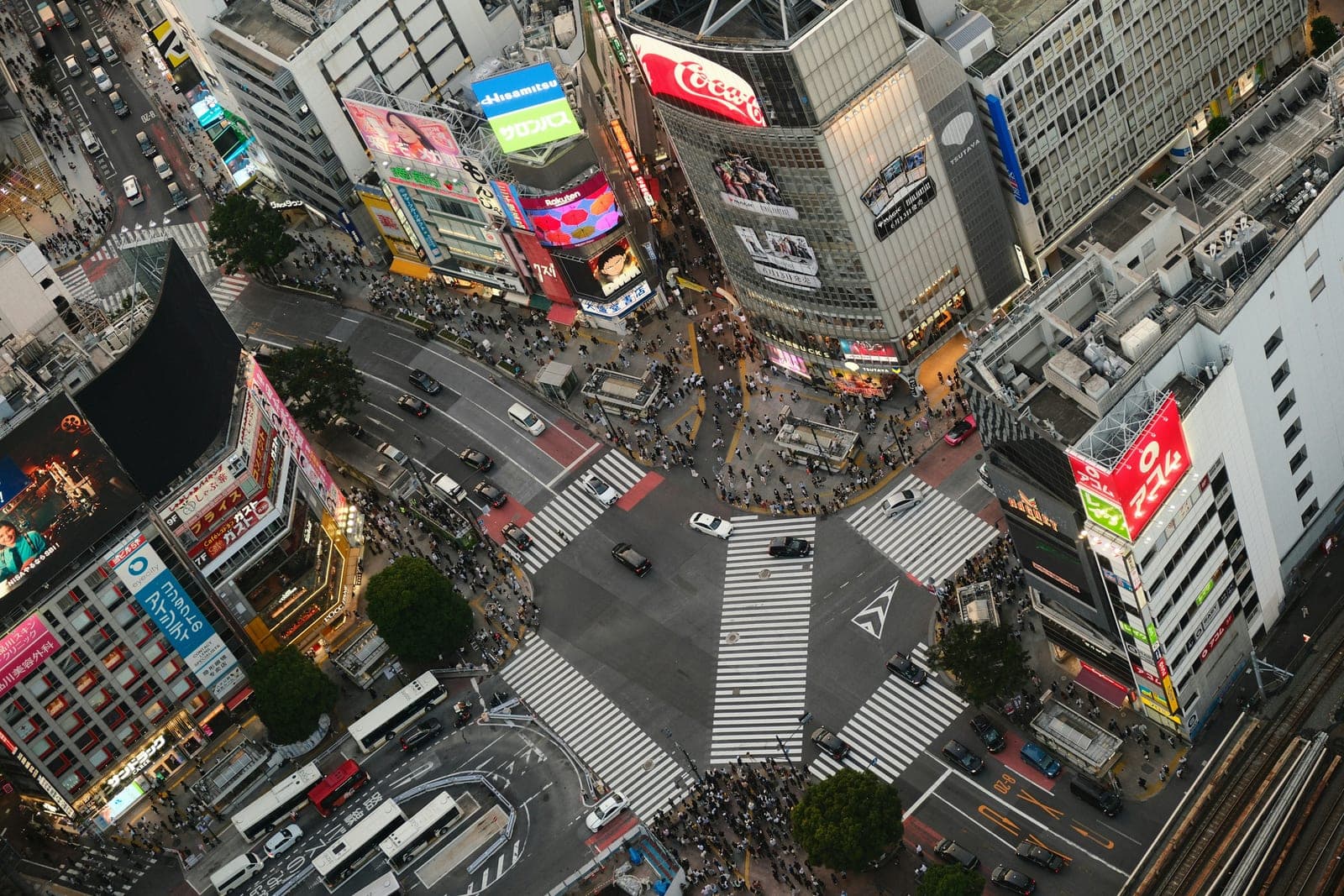 Vista aérea del cruce de Shibuya al anochecer desde arriba, con los pasos peatonales pintados en el pavimento y las pantallas publicitarias ya encendidas