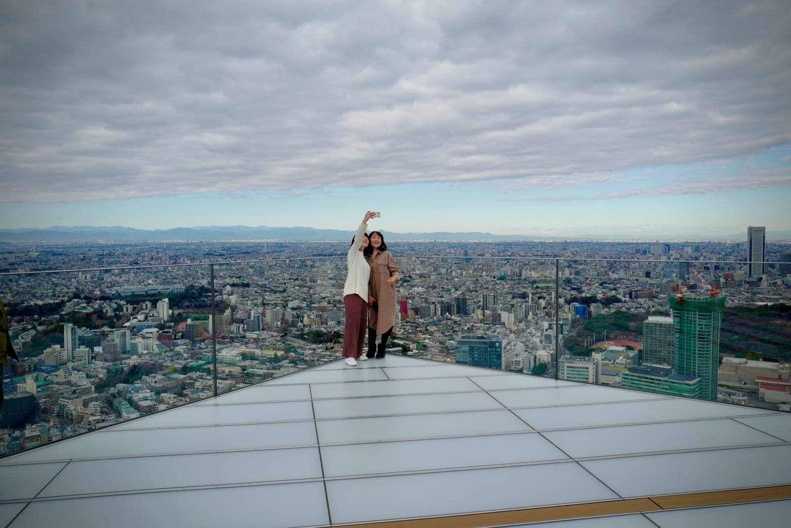 Dos visitantes tomándose una selfie en la punta de la terraza al aire libre de Shibuya Sky, con el horizonte de Tokio y montañas al fondo bajo un cielo nublado