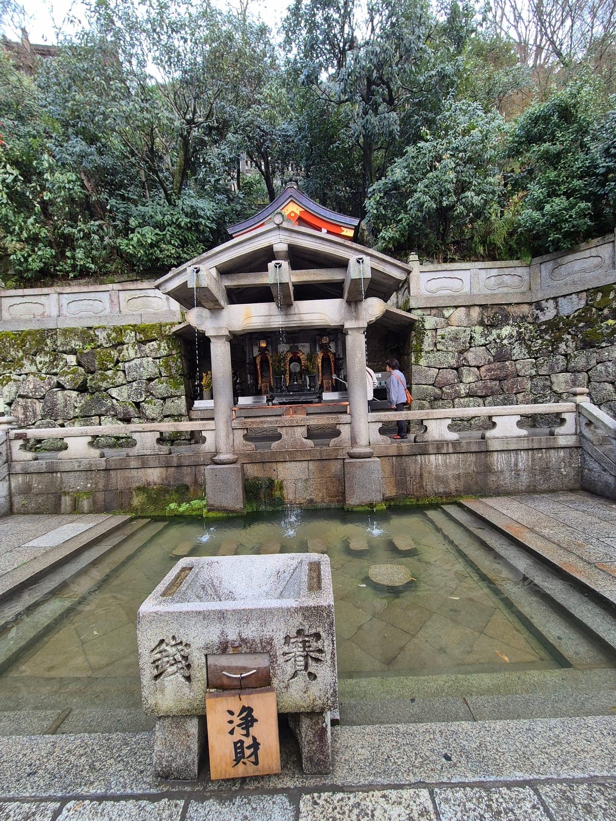 La fuente Otowa-no-taki del templo Kiyomizu-dera, con sus tres caños de agua bajo un techo de madera junto a un pequeño santuario