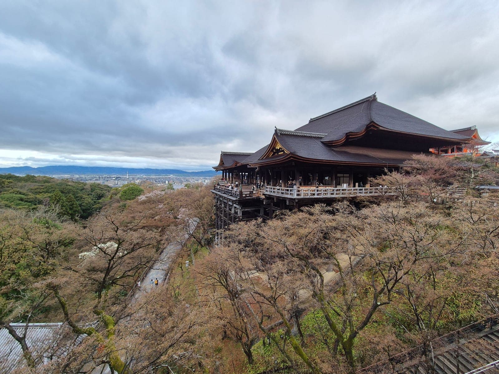 La terraza principal de Kiyomizu-dera con visitantes junto a la baranda de madera y la ciudad de Kioto y las montañas al fondo