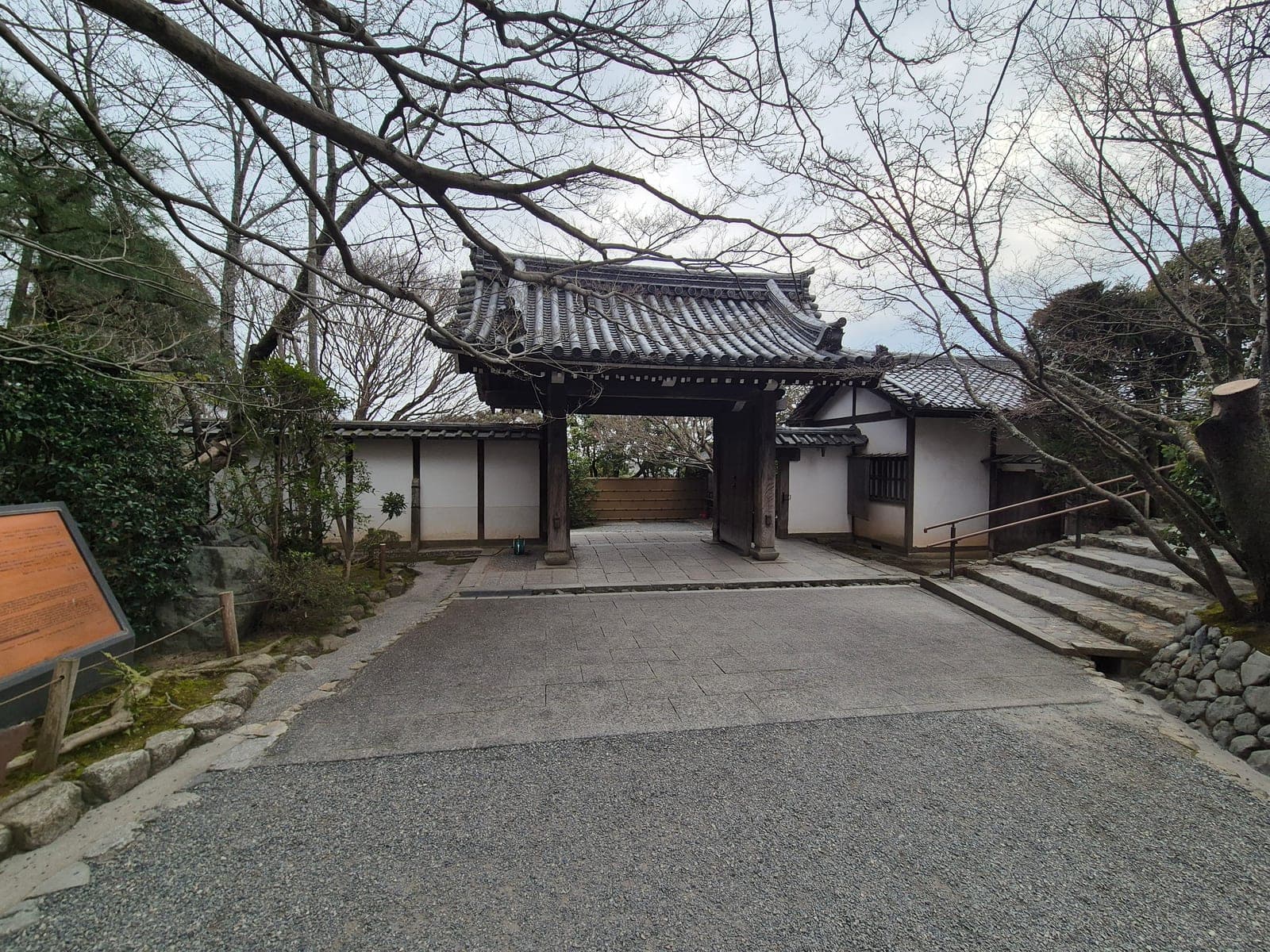 La puerta de acceso al recinto del templo Ryōan-ji en Kioto, entre árboles sin hojas, con un letrero informativo junto al camino