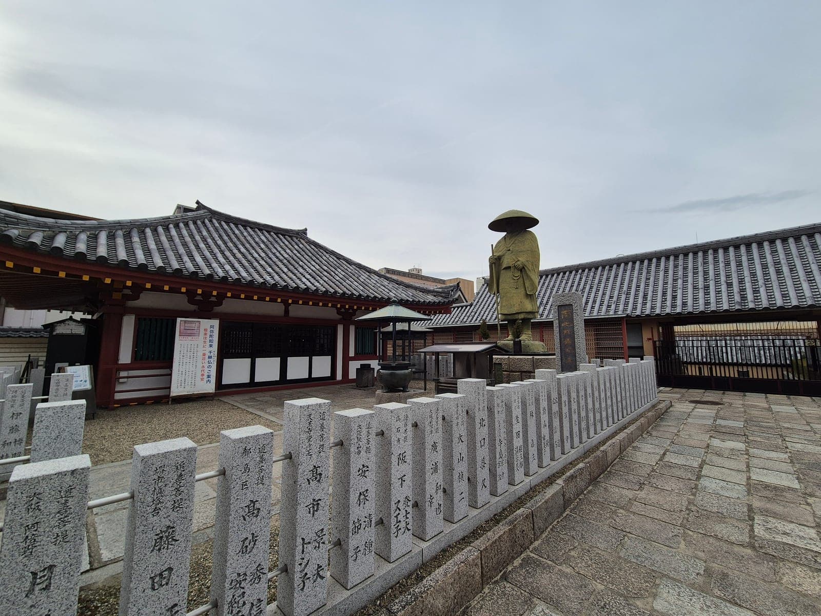 Estatua de bronce de un peregrino budista con sombrero cónico y bastón, rodeada de hileras de estelas de piedra conmemorativas en el recinto del templo Shitennōji