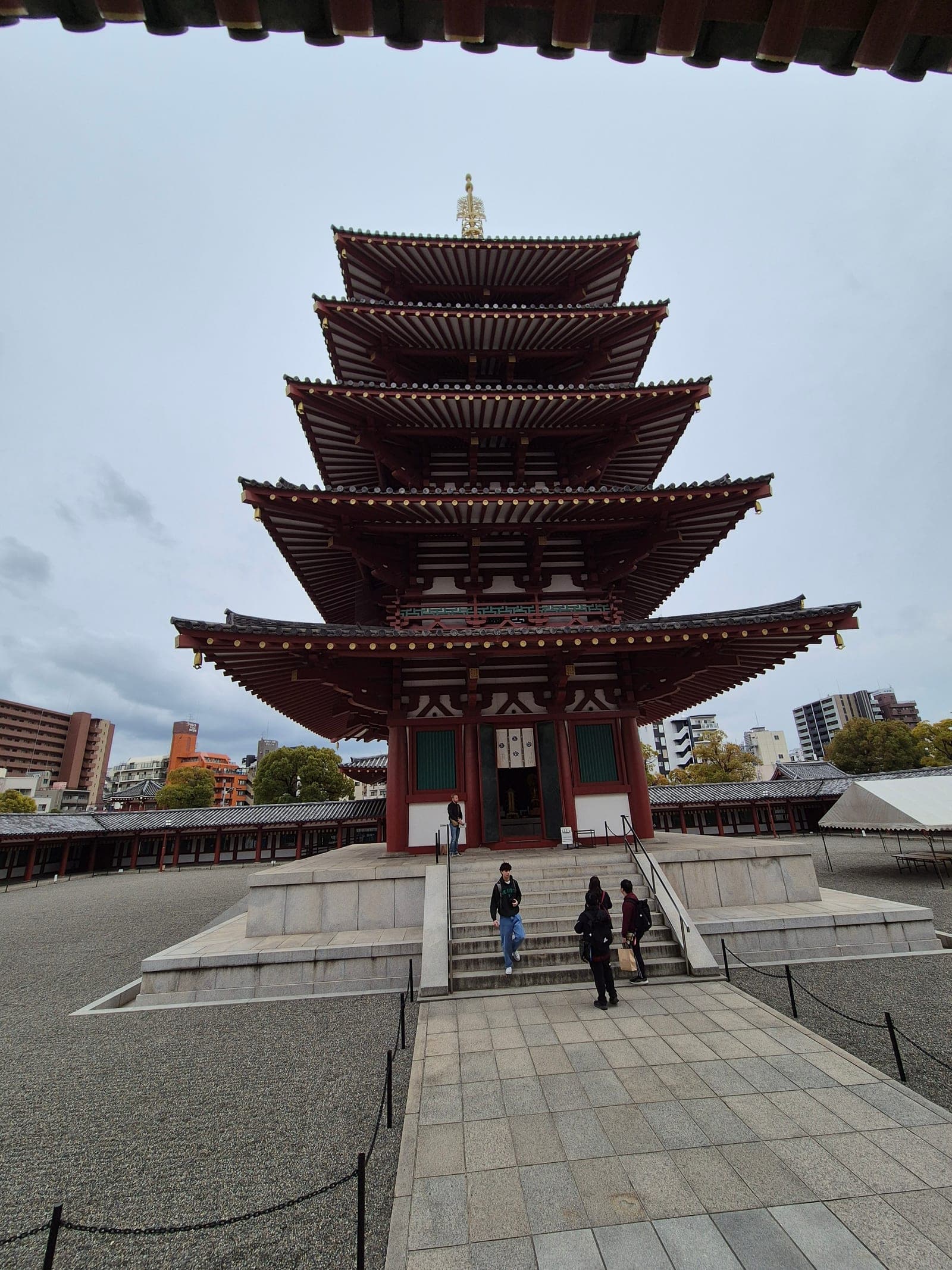 La pagoda de cinco pisos del templo Shitennōji vista de frente desde la escalinata de acceso, con visitantes bajando los escalones bajo un cielo nublado