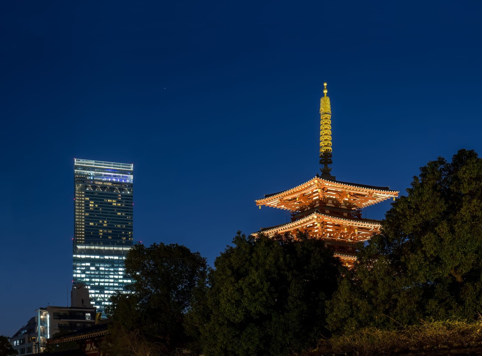 La pagoda de cinco pisos del Shitennōji iluminada al anochecer, con su remate dorado brillando y el rascacielos Abeno Harukas encendido al fondo
