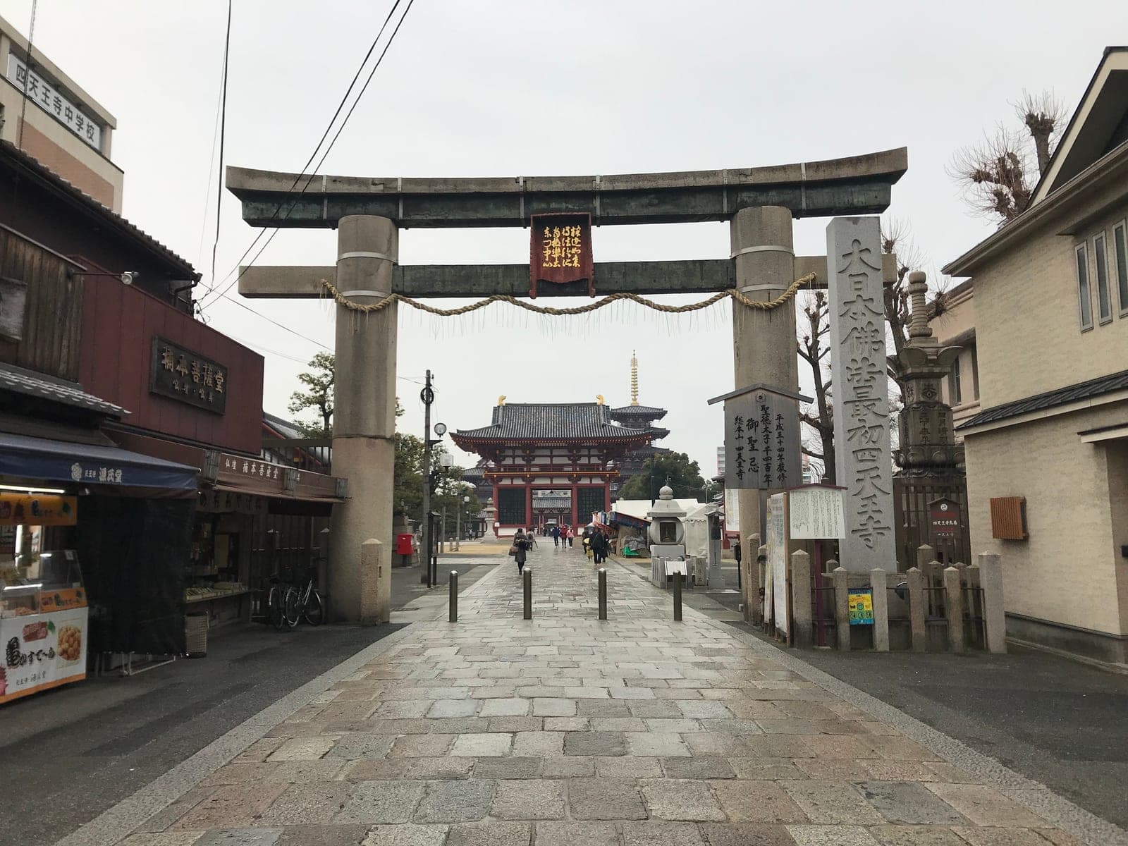 El gran torii de piedra del Shitennōji en la entrada oeste, con su placa central, la cuerda shimenawa y la puerta y la pagoda del templo al fondo de la calle