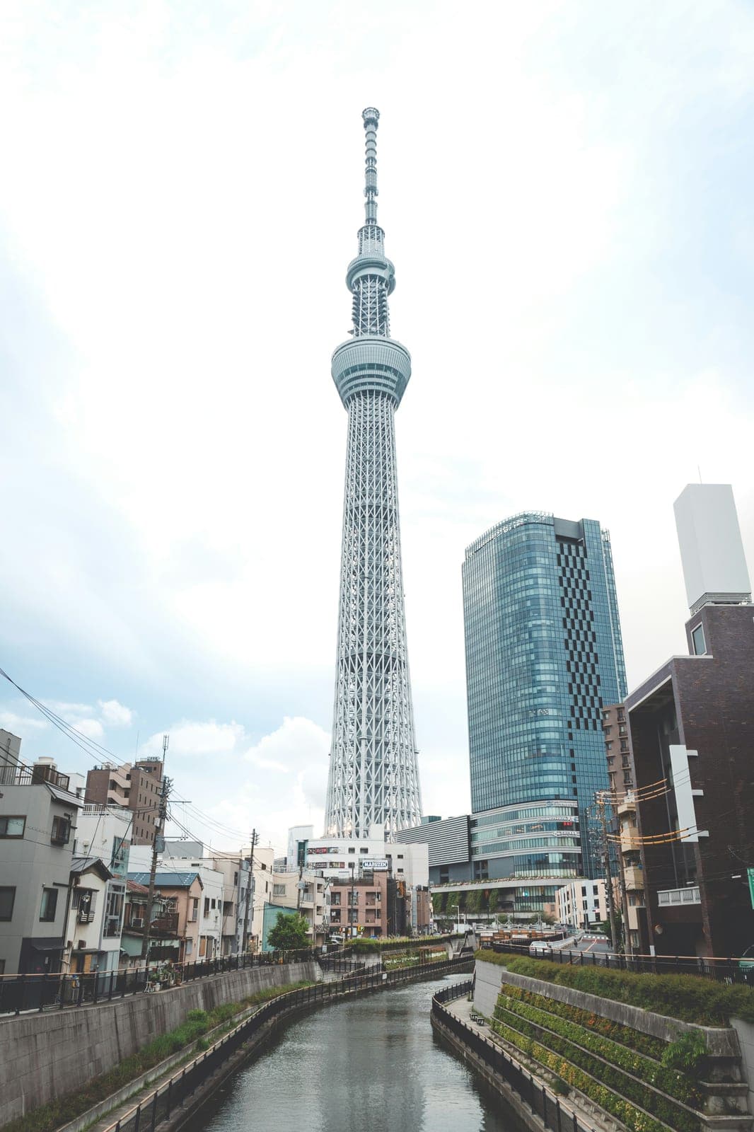 La Tokyo Skytree de cuerpo completo en un día nublado, vista desde un canal en Sumida, con la aguja de transmisión en la punta y edificios alrededor