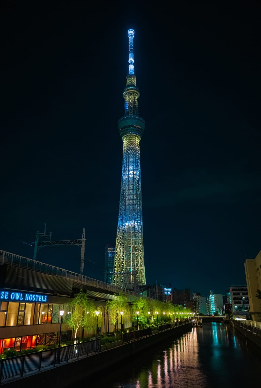 La Tokyo Skytree iluminada de azul y blanco de noche, reflejada en un canal de Sumida, con letreros de neón y árboles iluminados en la orilla